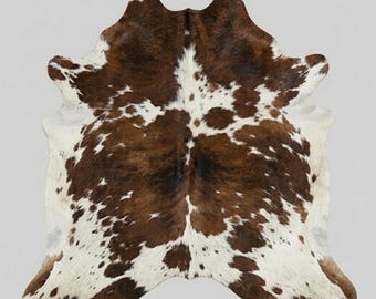 Puede incluir: Una alfombra de piel de vaca con un patrón natural de manchas irregulares marrones y blancas. La piel tiene una forma orgánica distintiva con un área central de color marrón oscuro que se extiende hacia manchas más pequeñas y secciones blancas.