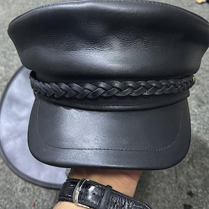 A black leather military-style cap featuring a smooth crown and a structured visor. A braided leather band wraps horizontally around the base of the crown, secured with stitching. The cap has a matte finish and a classic flat-top design.