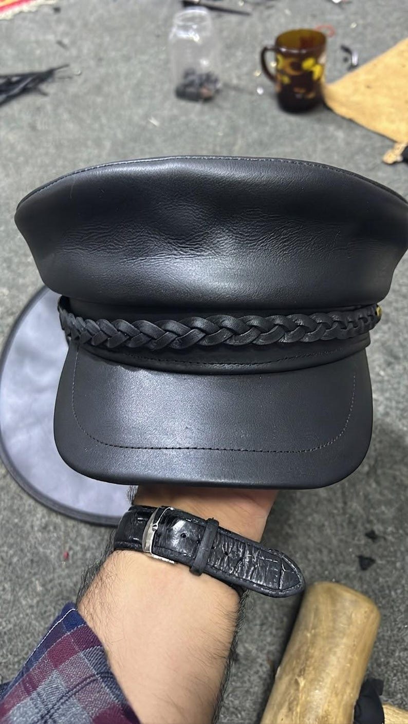 A black leather military-style cap featuring a smooth crown and a structured visor. A braided leather band wraps horizontally around the base of the crown, secured with stitching. The cap has a matte finish and a classic flat-top design.