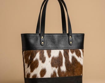 Peut inclure: Un sac cabas doté d'un panneau central en cuir de vachette poilu marron et blanc, bordé de cuir noir lisse en haut et en bas. Le sac comprend deux anses en cuir noir fixées par des rivets métalliques dorés sur le bord supérieur du sac.