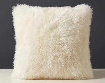 Peut inclure: Un coussin carré recouvert d'une longue fourrure synthétique ébouriffée de couleur blanc cassé. La texture est dense et duveteuse, créant un aspect doux sur toute la surface du coussin. L'objet conserve une forme uniforme, moelleuse et volumineuse.