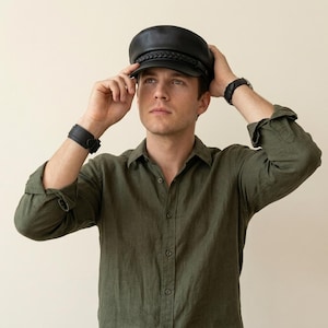 A black leather fiddler newsboy cap featuring a structured crown, a short stiff brim, and a decorative braided leather band across the front. The cap has a smooth finish and a classic rounded shape with a flat top panel and a curved visor.