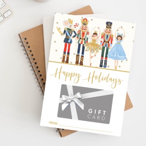 May include: A white gift card with a gray background and a white bow. The card has the text "Happy Holidays" and "Gift Card" printed on it. The card features a watercolor illustration of a nutcracker, a mouse, and two ballerinas.