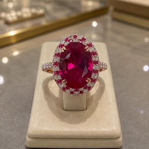 Peut inclure: Une bague ornée d'une grande pierre centrale rose foncé taille ovale, entourée d'un double halo de petites pierres roses et blanches. L'anneau est en métal et est serti de pierres blanches supplémentaires le long de la tige visible.