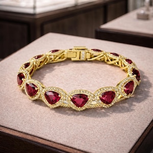 Può includere: Un bracciale dorato con pietre preziose triangolari rosso intenso e piccoli accenti trasparenti. Il bracciale è esposto su una superficie dai toni neutri, che ne mette in risalto il design intricato e l'appeal lussuoso. Anche la chiusura è dorata.