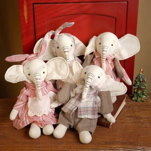 May include: Four handmade elephant plush toys. The elephants have white bodies, large ears, and long trunks. They are dressed in red and white gingham or plaid outfits, with some wearing aprons or pants. A small Christmas tree is in the background.