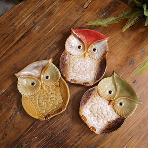 May include: Three ceramic owl-shaped dishes arranged on a wooden surface. Each dish features a distinct color scheme: one with a red top, one with a yellow-brown finish, and one with a green top. All three have textured feather patterns.