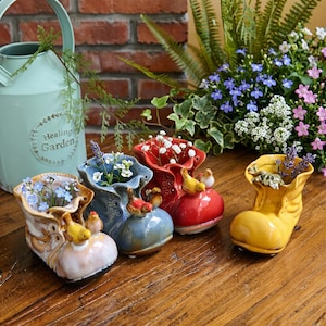 May include: Four ceramic shoe-shaped planters in light pink, blue, red, and yellow, each featuring small bird figurines perched on the front and filled with various small flowers, arranged on a wooden surface next to a light green metal watering can.