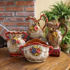 Peut inclure: Un ensemble de quatre récipients en céramique ornés de décorations florales en relief dans des tons de rouge, jaune, bleu et vert. Le lot comprend deux pichets avec anses et deux bols à bords festonnés, tous dotés d'un vernis brillant nuancé.