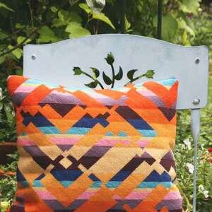 Puede incluir: Un coj&iacute;n de decoraci&oacute;n geom&eacute;trico de colores con rayas naranjas, rosas, azules y marrones. El coj&iacute;n est&aacute; sentado en una silla de metal blanca.