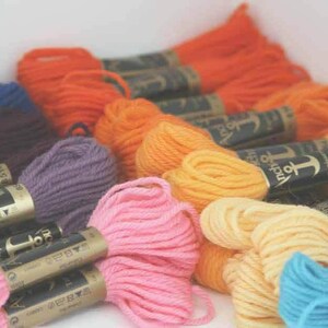 Puede incluir: Surtido de carretes de hilo de bordar de la marca Anchor en varios colores, incluyendo morado, rosa, naranja, amarillo y azul. Cada carrete tiene una etiqueta dorada con el logotipo y el texto de Anchor.