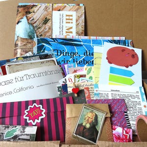 Könnte beinhalten: Ein Pappkarton gefüllt mit verschiedenen Papier-Ephemera, darunter Postkarten, Umschläge und Aufkleber. Sichtbarer Text umfasst "Hi Mom," "Dinge, die wir lieben," "Oase für Traumtänzer" und "Venice, California." Ein Klebeband und eine Briefmarke liegen bei.
