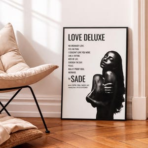 May include: A black and white framed poster featuring the album Love Deluxe by Sade. The print displays the title, tracklist, artist name, genre, and running time alongside a photographic portrait of a person with long hair, arms crossed.