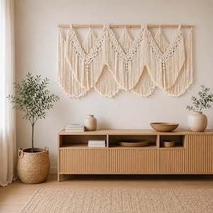 May include: A large off-white macrame wall hanging featuring intricate knotted patterns and long fringe, suspended from a horizontal wooden rod. The piece displays a series of V-shaped sections with detailed diamond-shaped knots and vertical cords.