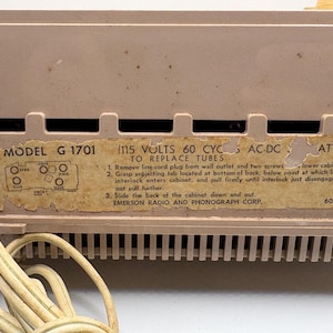 May include: Close-up of a vintage Emerson radio. The radio is a light tan color with a label that reads "MODEL G 1701". The label also includes instructions for replacing tubes. A power cord is visible.