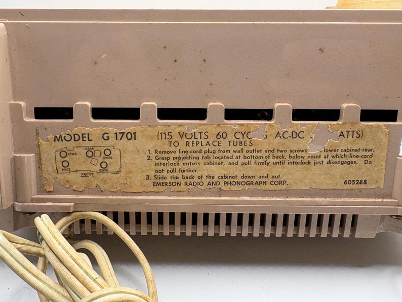 May include: Close-up of a vintage Emerson radio. The radio is a light tan color with a label that reads "MODEL G 1701". The label also includes instructions for replacing tubes. A power cord is visible.