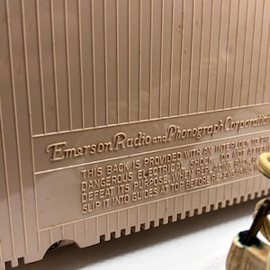 May include: Close-up of a vintage Emerson Radio and Phonograph Corporation device. The beige casing has vertical ridges and a rectangular panel with embossed text. The text warns about electrical shock and proper replacement of the back.