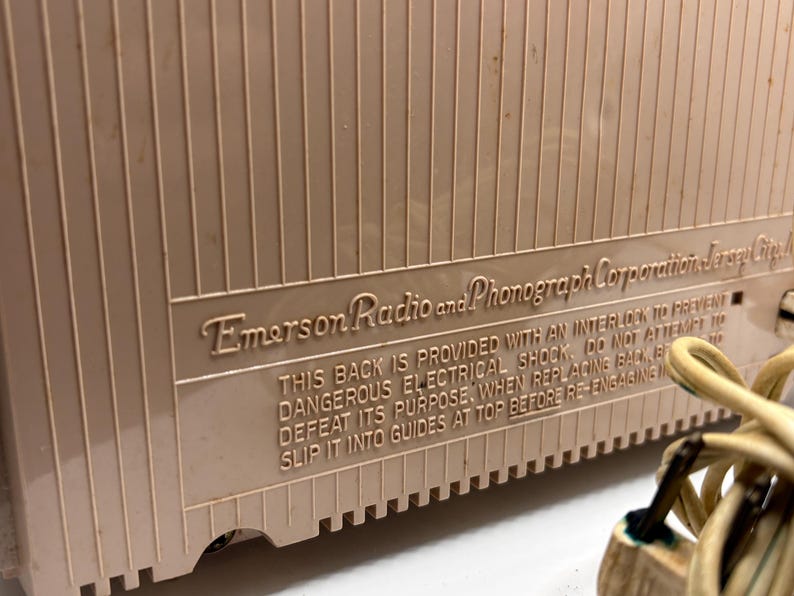 May include: Close-up of a vintage Emerson Radio and Phonograph Corporation device. The beige casing has vertical ridges and a rectangular panel with embossed text. The text warns about electrical shock and proper replacement of the back.