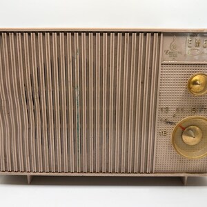 May include: Vintage Emerson radio in a light brown color. The radio has a vertical ribbed front panel and a control panel with two gold-colored knobs and a dial. The Emerson logo is visible on the top right corner. A power cord is attached to the radio.