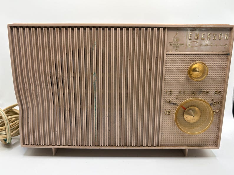 May include: Vintage Emerson radio in a light brown color. The radio has a vertical ribbed front panel and a control panel with two gold-colored knobs and a dial. The Emerson logo is visible on the top right corner. A power cord is attached to the radio.