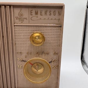 May include: Vintage Emerson Century radio with a tan and gold color scheme. The radio features a speaker grill, two gold knobs, and a tuning dial with a red indicator. The Emerson logo is visible on the top of the radio.