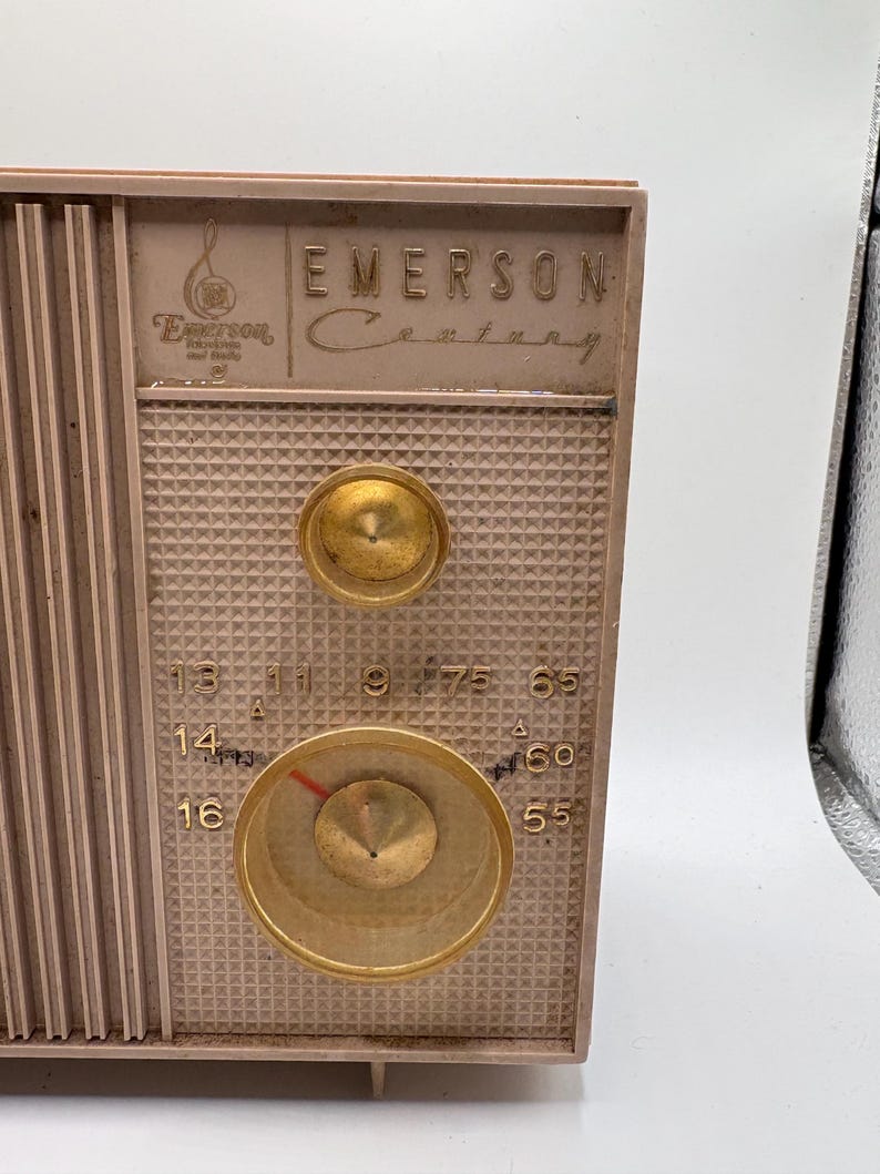 May include: Vintage Emerson Century radio with a tan and gold color scheme. The radio features a speaker grill, two gold knobs, and a tuning dial with a red indicator. The Emerson logo is visible on the top of the radio.