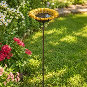 Puede incluir: Un ba&ntilde;o para p&aacute;jaros con un cuenco en forma de girasol amarillo con un centro texturizado lleno de agua, sostenido por una fina estaca de metal marr&oacute;n oscuro insertada en el suelo. El cuenco tiene bordes de p&eacute;talos detallados.