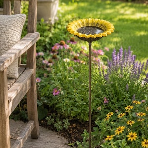 Puede incluir: Un ba&ntilde;o para p&aacute;jaros de jard&iacute;n con un cuenco en forma de girasol amarillo brillante con detalles centrales de color marr&oacute;n oscuro, montado sobre una estaca de metal fina y oscura. El cuenco contiene agua que refleja el entorno exterior.