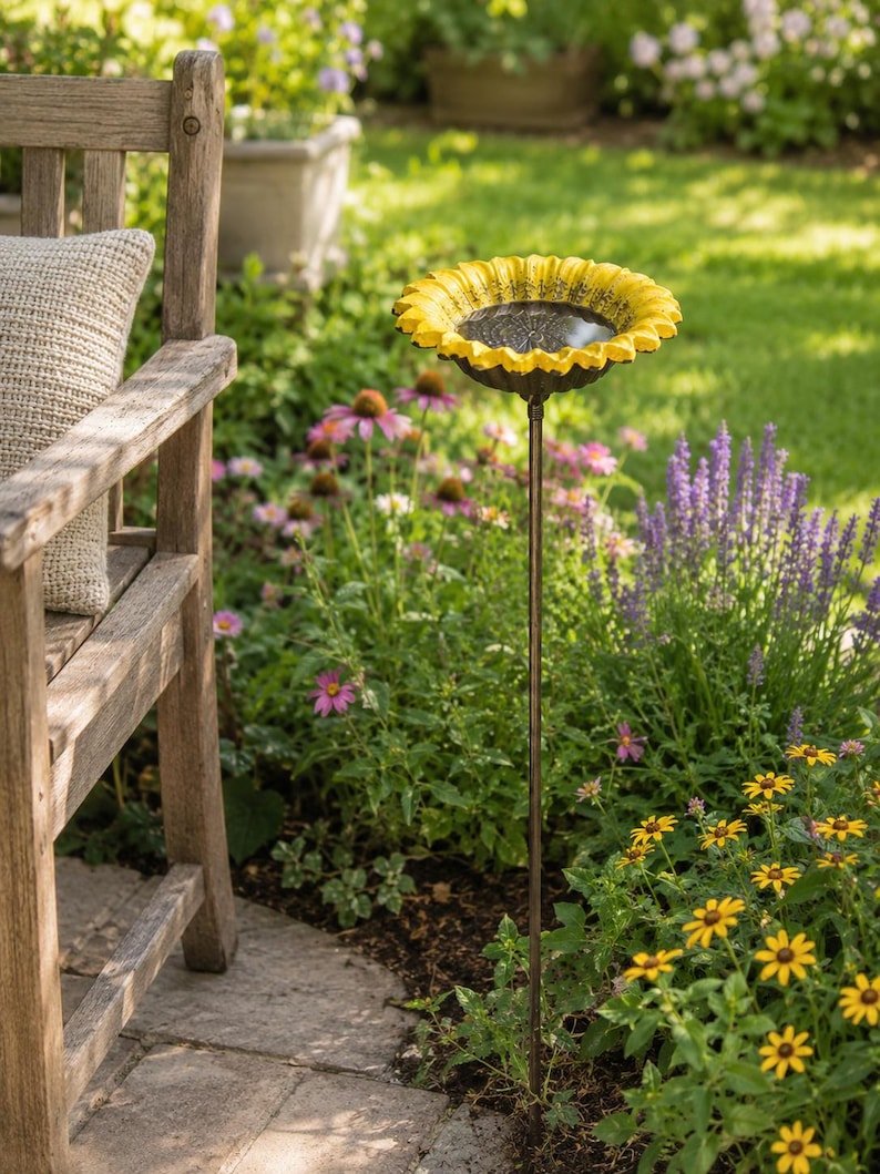 Puede incluir: Un ba&ntilde;o para p&aacute;jaros de jard&iacute;n con un cuenco en forma de girasol amarillo brillante con detalles centrales de color marr&oacute;n oscuro, montado sobre una estaca de metal fina y oscura. El cuenco contiene agua que refleja el entorno exterior.