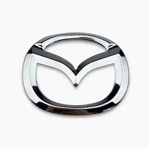May include: A chrome-finished Mazda vehicle emblem featuring the iconic stylized M logo enclosed within an oval frame. The emblem has a polished, reflective metallic surface with smooth, curved lines that form the central V-shaped design.