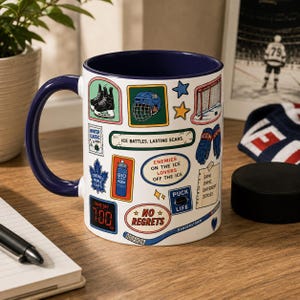 May include: A white ceramic mug with a dark blue interior and handle, featuring various ice hockey-themed stickers and text including "ICE BATTLES. LASTING SCARS.", "ENEMIES ON THE ICE LOVERS OFF THE ICE", "NO REGRETS", "PUCK LIFE", and "SAME GAME DIFFERENT JERSEY."