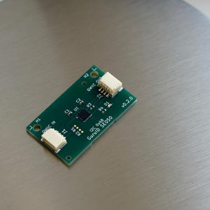 Puede incluir: Una pequeña placa de circuito verde rectangular con conectores blancos en cada extremo. La placa tiene varios componentes y texto, incluyendo "I2C 0x48 GurulD SE050 v0.2.0". La placa está sobre una superficie plateada.