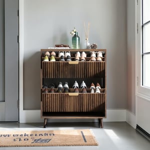 May include: A wooden shoe rack with three shelves, filled with various shoes. The rack has a ribbed design and gold-colored handles. A doormat with text is in front of the rack.