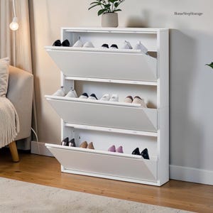May include: A white, three-tiered shoe storage cabinet with drop-down doors. Various shoes are visible inside the open compartments. A small potted plant sits on top. The text "HomeStepStorage" is in the upper right corner.