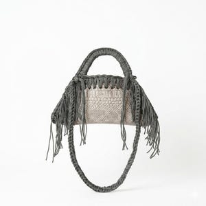 Puede incluir: Un bolso de mano gris con asa trenzada y flecos. El bolso presenta una sección central texturizada de color claro, y el diseño general es moderno y elegante. El bolso está diseñado para llevarse en la mano o al hombro.