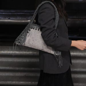 Puede incluir: Un bolso gris con estampado de piel de serpiente y flecos. El bolso tiene una correa trenzada y se lleva al hombro. El bolso tiene forma triangular.