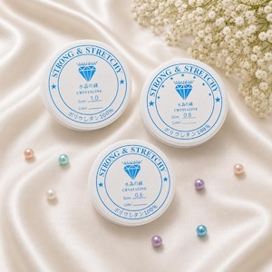 May include: Three white spools of strong and stretchy crystalline thread, labeled with sizes 1.0, 0.8, and 0.6. The spools have blue text and a diamond graphic. Scattered around are pastel-colored beads.