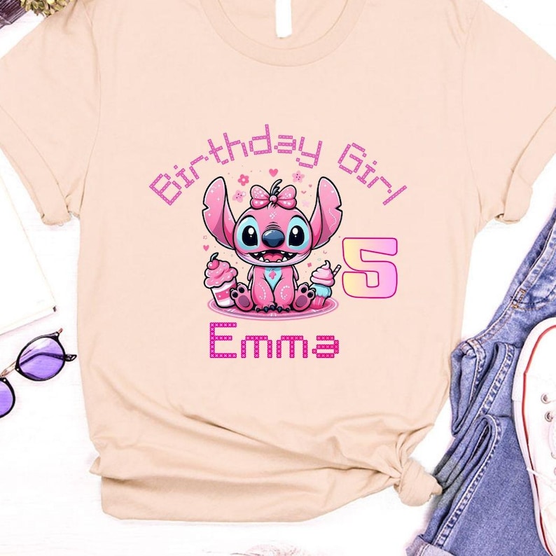 May include: A light beige t-shirt featuring a pink cartoon character with large ears, holding cupcakes. The text "Birthday Girl", the number "5", and the name "Emma" are also displayed in pink.