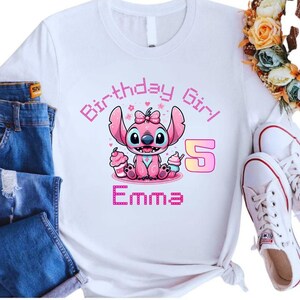 May include: White t-shirt featuring a pink cartoon character with the text "Birthday Girl" and the number "5". The character is holding cupcakes, and the name "Emma" is displayed below. The shirt is paired with blue jeans and white sneakers.