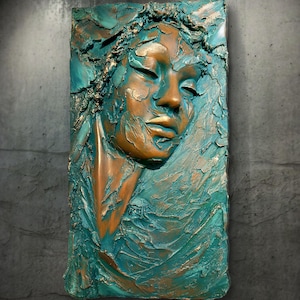 May include: A rectangular wall sculpture featuring a high-relief portrait of a face with closed eyes. The artwork is finished in a textured blend of metallic bronze and deep teal paint, creating a layered, weathered appearance across the surface.