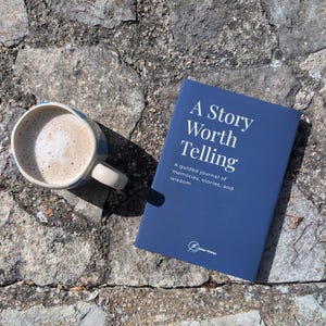 May include: A dark blue journal titled A Story Worth Telling: A guided journal of memories, stories, and wisdom with the Baker Press logo at the bottom, positioned next to a ceramic mug filled with a light brown coffee beverage with foam.