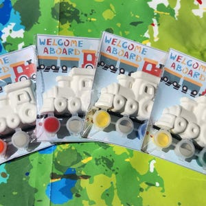 May include: Four white ceramic train engine figurines with the words "Welcome Aboard" printed on the packaging. Each figurine comes with four small paint pots in different colors.