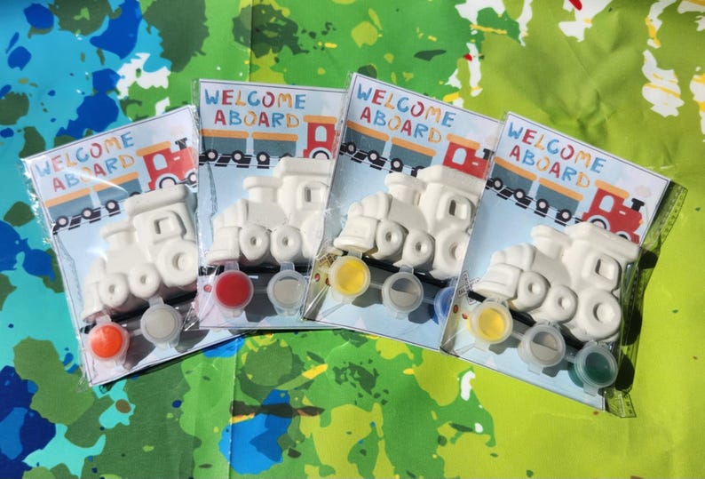 May include: Four white ceramic train engine figurines with the words "Welcome Aboard" printed on the packaging. Each figurine comes with four small paint pots in different colors.