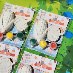 Peut inclure: Trois ensembles de kits de bricolage sur le th&egrave;me de l'automne &agrave; peindre soi-m&ecirc;me. Chaque kit comprend un &eacute;pi de ma&iuml;s, une citrouille et quatre petits pots de peinture. Les kits sont emball&eacute;s dans des sacs en plastique transparent avec une carte qui dit "Happy Fall Y'all".