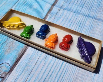 Puede incluir: Un juego de seis crayones de colores con forma de naves espaciales y platillos voladores. Los crayones están dispuestos en fila en una caja de cartón.