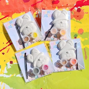 Puede incluir: Cuatro kits de pintura con forma de conejito de Pascua. Cada kit incluye una figura de conejito blanco, pequeños botes de pintura de varios colores y un pincel. Los kits están empaquetados en plástico transparente con "HAPPY EASTER" impreso.