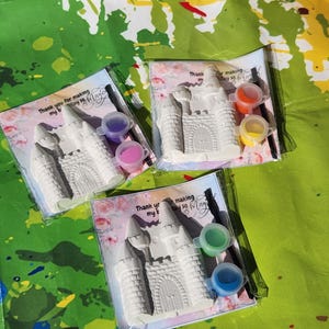 May include: Three castle-shaped paint kits in clear plastic packaging. Each kit includes a white castle, small paint pots in various colors, and a paintbrush. The packaging has text that reads "Thank you for making my birthday so Magic".