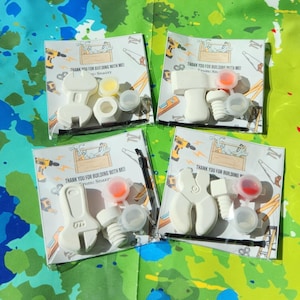 May include: Four favour boxes with a tool theme. Each box contains a white plastic tool, a small bottle of paint, and a small plastic lid. The boxes have a printed label with the text "Thank you for building with me" and "From Snazzy".