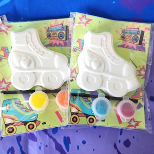 May include: Two white ceramic roller skate paint-by-number kits with paint pots in yellow, orange, blue, and red. The kits feature a retro design with a boombox and stars.