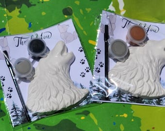 May include: Two paint-your-own wolf head kits. Each kit includes a white ceramic wolf head, paint pots, and a paintbrush. The packaging features paw prints and the words "Thank You". The background is a colourful, abstract design.